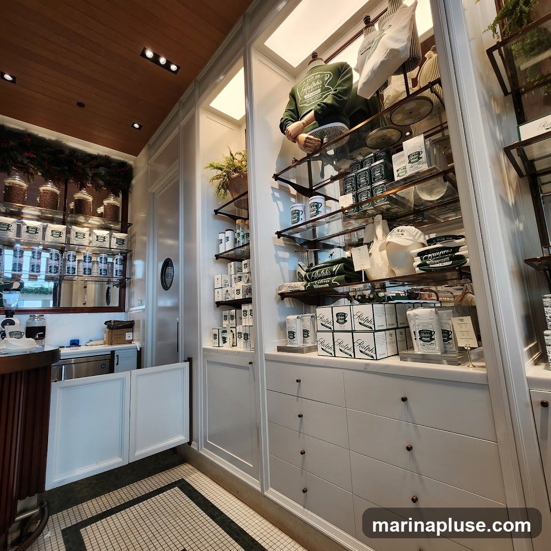 A well-lit interior display of Ralph's Coffee branded merchandise. Shelves are stocked with various items including mugs, hats, bags, and packaged goods. A mannequin is dressed in a green sweatshirt with the Ralph's Coffee logo. The display features a clean, organized aesthetic with white shelving and drawers.