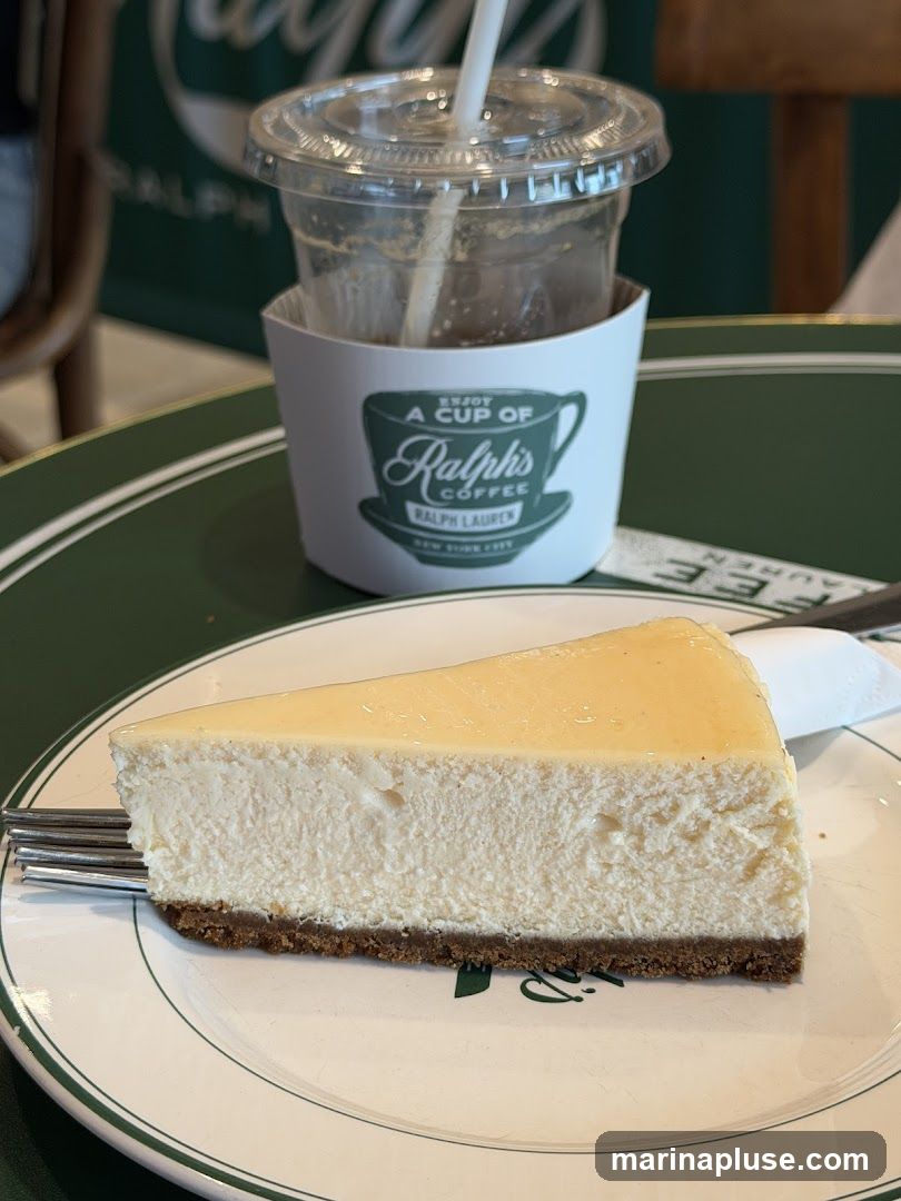 A slice of cheesecake sits on a white plate with a green rim. A fork rests on the left side of the plate. In the background, a clear plastic cup with a straw and a white coffee sleeve is visible. The coffee sleeve has a logo that says "ENJOY A CUP OF Ralph's COFFEE RALPH LAUREN NEW YORK CITY". The table surface is green with white stripes.