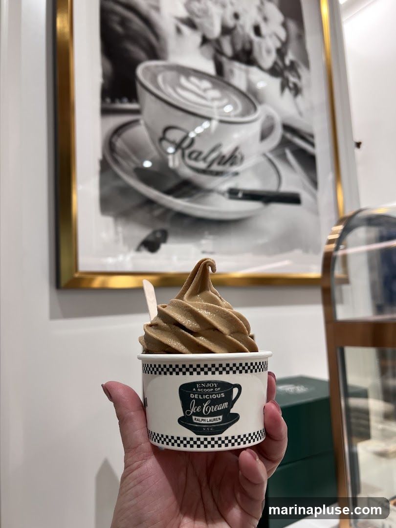 A hand holds a cup of soft-serve ice cream. The cup has a black and white checkered border and a graphic of a coffee cup with the words "Enjoy a scoop of delicious Ice Cream Ralph Lauren N.Y.C." The background features a framed black and white photograph of a coffee cup with latte art.