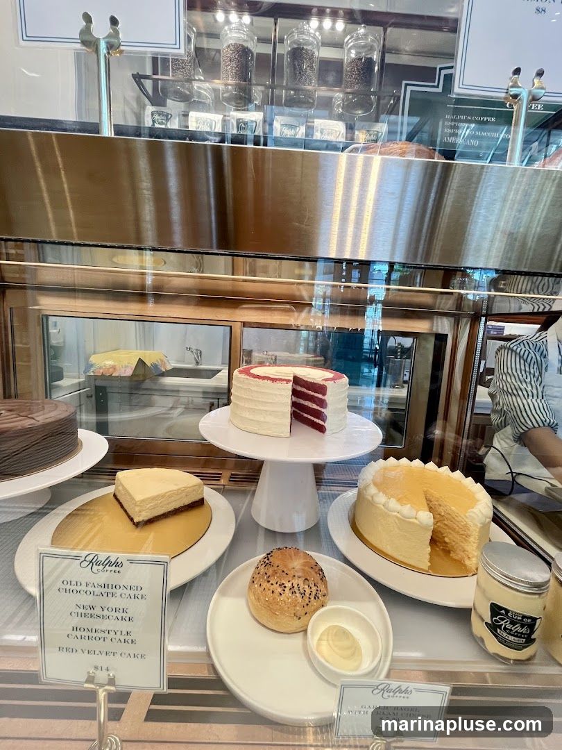 A display case filled with various cakes and pastries. Visible items include a red velvet cake, a New York cheesecake, a chocolate cake, and a garlic bagel with cream cheese. There are also jars of coffee beans and branded coffee cups.