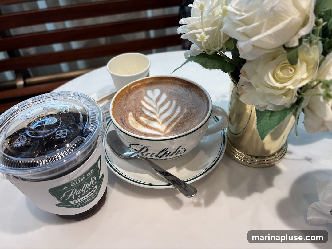 A close-up shot of two coffee drinks on a white table. In the foreground, a clear plastic cup with a lid contains a dark beverage, possibly iced coffee. A paper sleeve around the cup has a green logo that reads "A Cup of Ralph's Coffee New York City". To the right, a white ceramic mug with a saucer holds a latte with intricate foam art. The mug and saucer have a green rim and the word "Ralph's" is visible on the mug. A silver spoon rests on the saucer. In the background, a vase filled with white roses and greenery sits on the table. A small white paper cup is also visible. The table is set against a backdrop of dark wooden slatted seating.