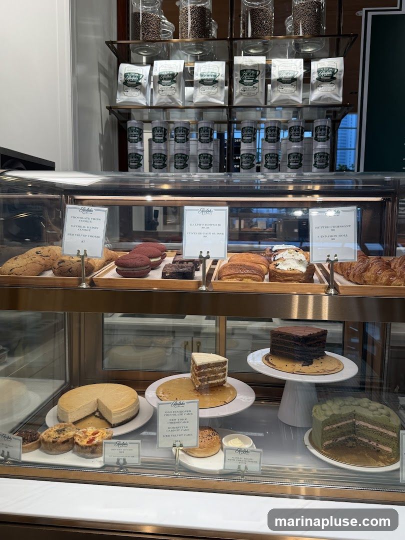 A display case filled with various pastries, cakes, and cookies. On the top shelf, bags of coffee beans and packaged coffee are visible. The display case features items like chocolate chip cookies, red velvet macarons, brownies, croissants, cheesecakes, carrot cake, chocolate cake, and a green layered cake. Labels indicate some of the items and their prices.
