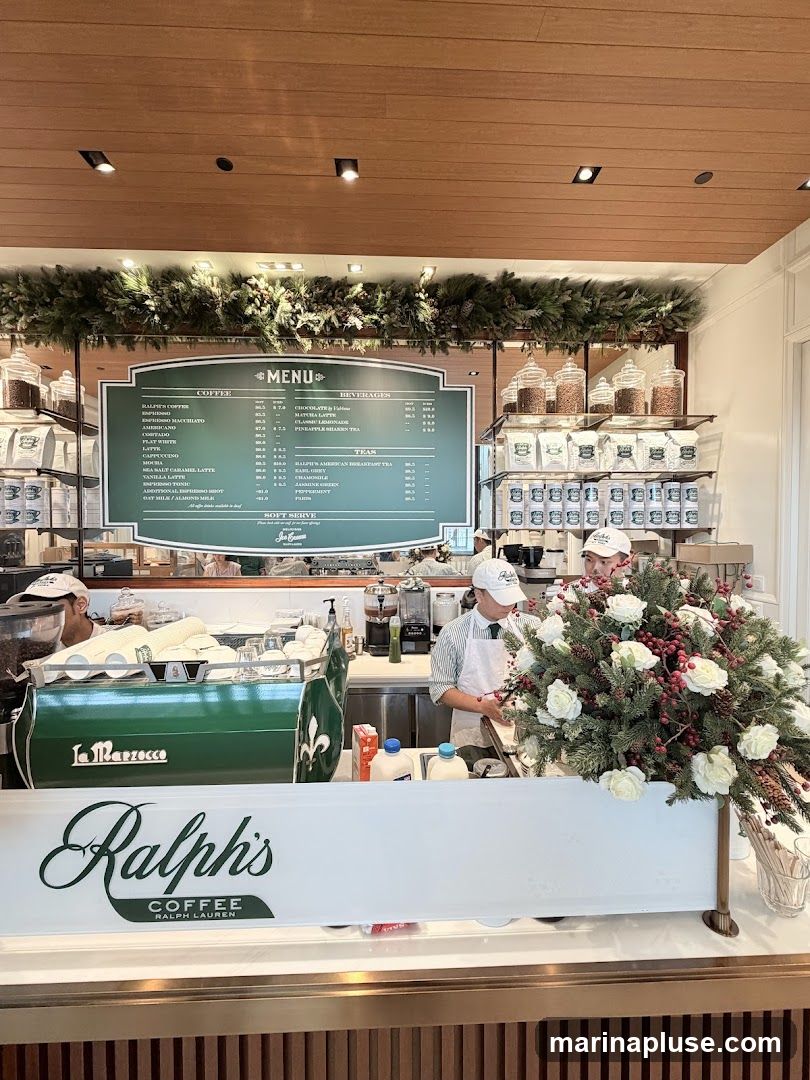 A view of the counter at Ralph's Coffee, with a menu board displayed above. Baristas are visible behind the counter, and a festive floral arrangement is in the foreground. Jars of coffee beans and packaged coffee are on shelves.
