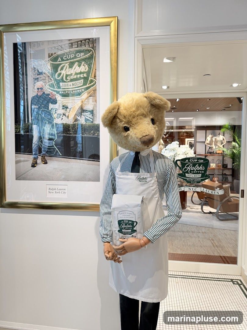 A mannequin dressed as a barista, wearing a striped shirt, tie, and apron, holds a bag of Ralph's Coffee. Behind the mannequin is a framed photograph of Ralph Lauren in front of a Ralph's Coffee storefront. The background shows the interior of a Ralph Lauren store with clothing displays.