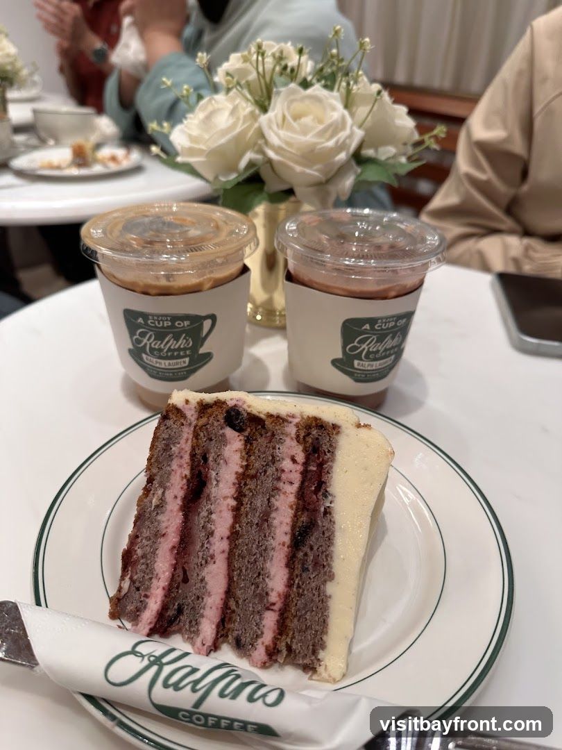 A slice of layered cake with pink frosting and berries sits on a white plate with a green rim. Two iced coffee drinks in clear cups with branded sleeves are behind the cake. The sleeves have the Ralph's Coffee logo. A bouquet of white roses is in a gold vase in the background. People are seated at tables in the background.