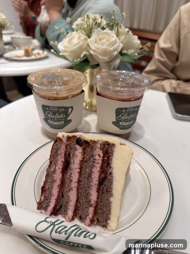 A slice of layered cake with pink frosting and berries sits on a white plate with a green rim. Two iced coffee drinks in clear cups with branded sleeves are behind the cake. The sleeves have the Ralph's Coffee logo. A bouquet of white roses is in a gold vase in the background. People are seated at tables in the background.