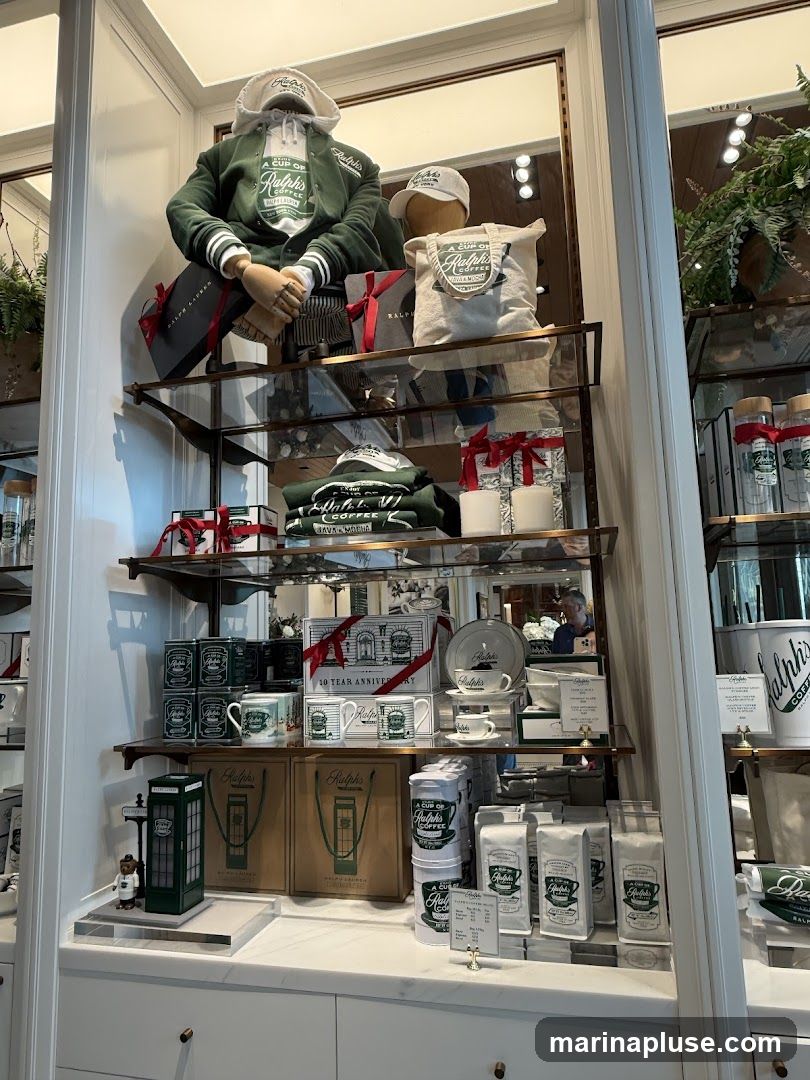 A display of Ralph Lauren branded coffee merchandise, including mugs, coffee tins, bags of coffee beans, and tote bags. Two mannequins are dressed in Ralph Lauren apparel, one wearing a varsity jacket and the other a hoodie. Gift-wrapped boxes and decorative plants are also visible.