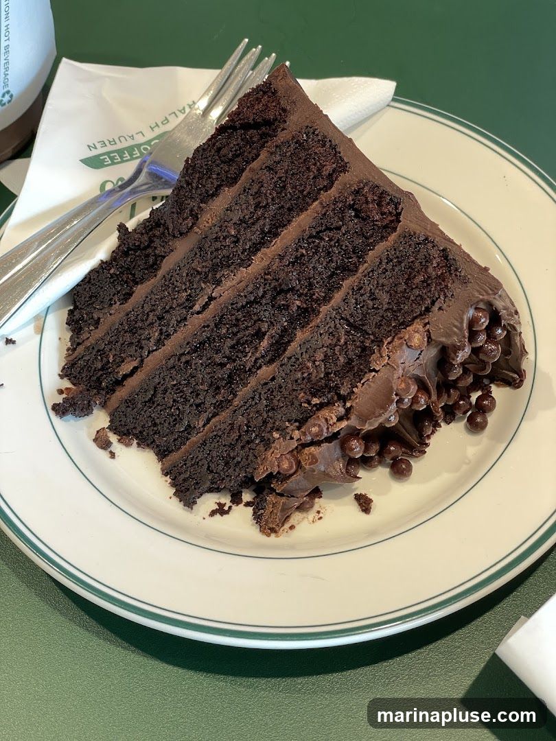 A slice of rich chocolate cake with multiple layers of dark chocolate cake and chocolate frosting. The cake is topped with chocolate frosting and chocolate pearls. It is served on a white plate with a green rim, alongside a fork and a napkin with the Ralph Lauren Coffee logo.