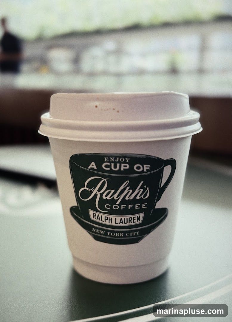 A close-up shot of a white paper coffee cup with a dark green logo for "Ralph's Coffee" by Ralph Lauren, New York City. The cup has a white lid and appears to be filled with a beverage. The background is blurred, showing a person in silhouette and what looks like a counter or bar area.
