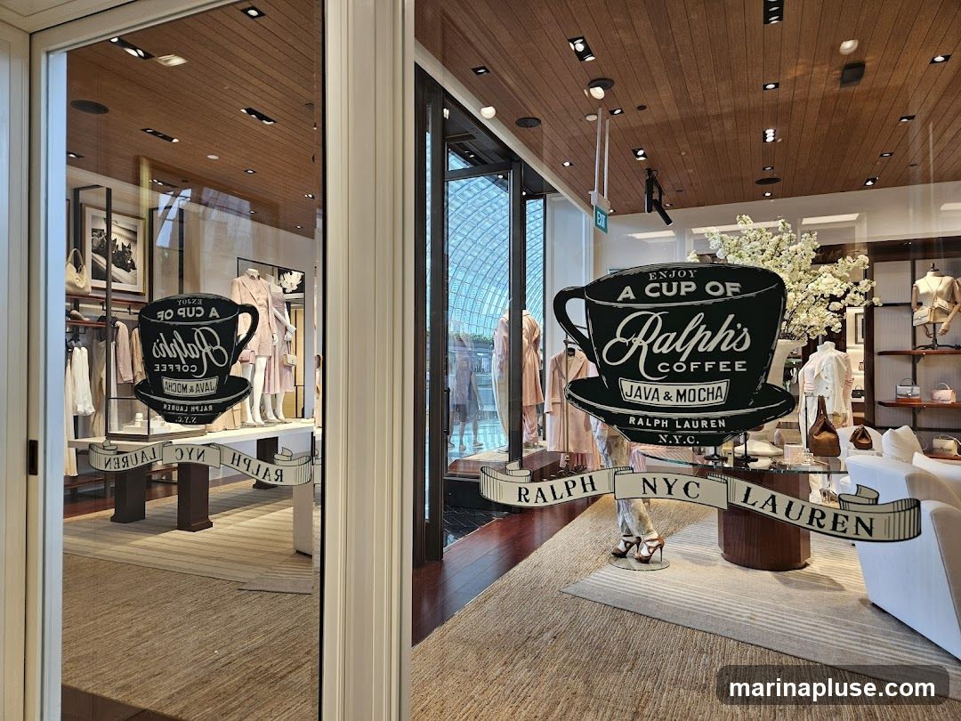 A window display for Ralph's Coffee, featuring a large graphic of a coffee cup and saucer with the text "ENJOY A CUP OF Ralph's COFFEE JAVA & MOCHA RALPH LAUREN N.Y.C.". A banner below reads "RALPH NYC LAUREN". The display is set against a backdrop of clothing racks with apparel and mannequins, suggesting it is part of a Ralph Lauren store.
