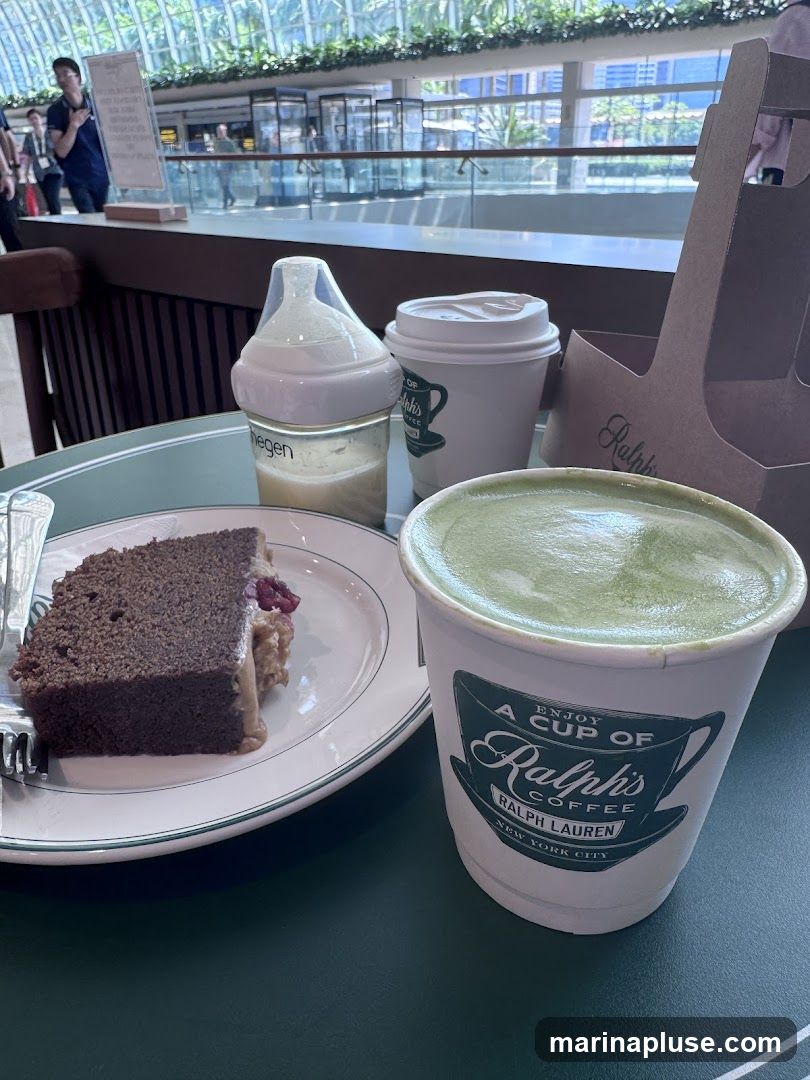 A slice of chocolate cake with a creamy filling and berries sits on a plate next to a cup of matcha latte. A baby bottle and another coffee cup are also on the table. The cups and a paper carrier have the Ralph's Coffee logo.