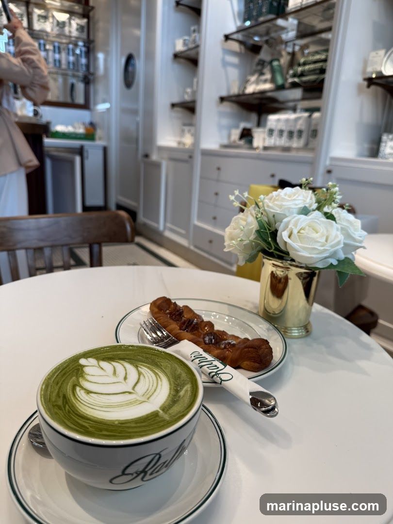 A close-up shot of a matcha latte with intricate foam art and a pastry on a white table. The latte is in a white cup with green trim, and the pastry appears to be a twisted croissant with nuts and glaze. A white napkin with a logo is placed next to the pastry. In the background, shelves are stocked with various items, and a vase of white roses adds a decorative touch.