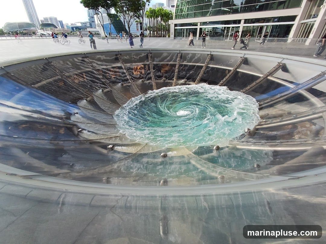A close-up view of a water vortex fountain in an urban setting. The water is swirling in a circular motion, creating a mesmerizing effect. In the background, people are seen walking and cycling along a promenade, with modern buildings and skyscrapers visible. The fountain appears to be made of metal and glass, with a sleek, contemporary design.
