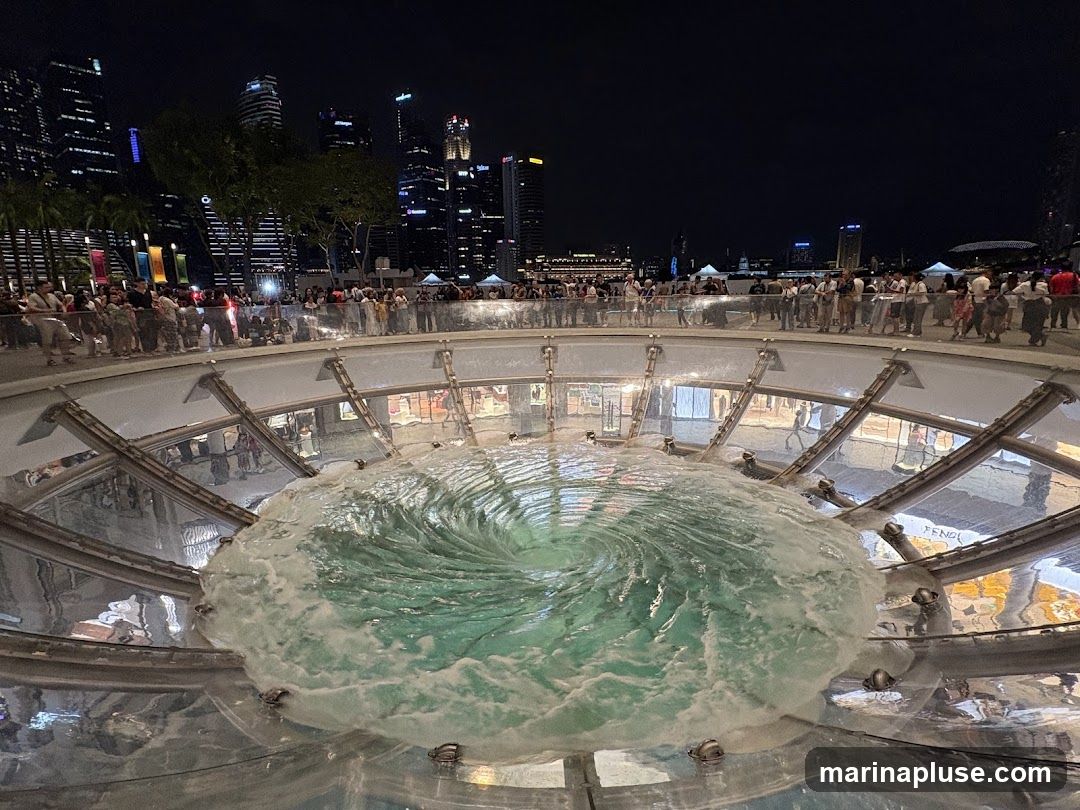 A large, circular fountain with water swirling into a vortex is illuminated at night. People are gathered around the edge of the fountain, observing the spectacle. In the background, the illuminated skyline of a city is visible, with tall buildings and lights. The scene appears to be in a public space or shopping area.