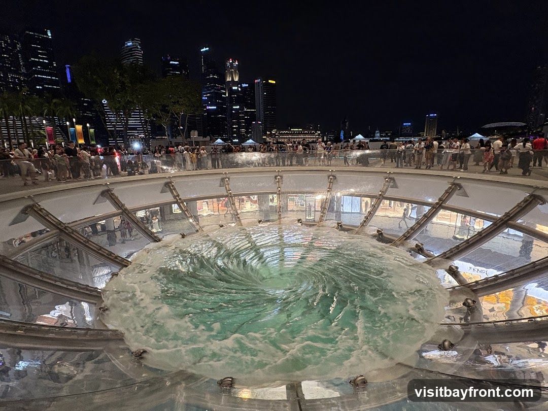 A large, circular fountain with water swirling into a vortex is illuminated at night. People are gathered around the edge of the fountain, observing the spectacle. In the background, the illuminated skyline of a city is visible, with tall buildings and lights. The scene appears to be in a public space or shopping area.