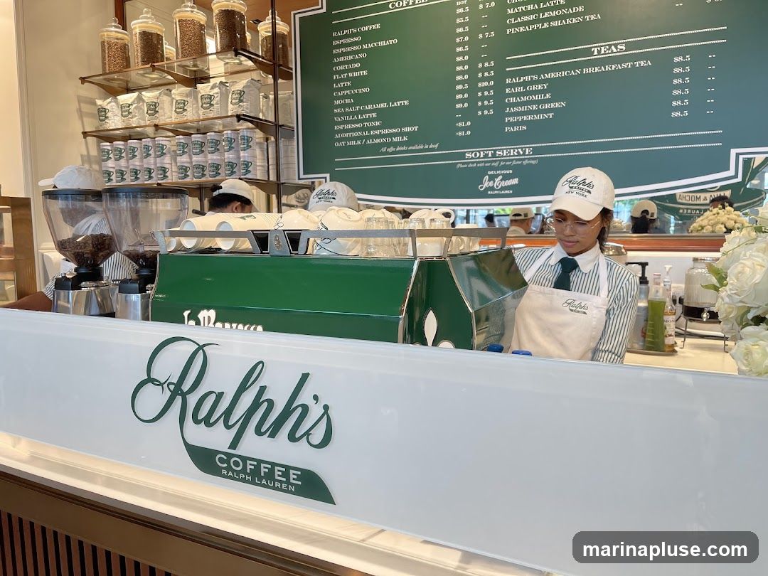 A view of a Ralph's Coffee counter with a barista in uniform. Behind the counter are shelves stocked with coffee beans, bags of coffee, and stacks of cups. A large menu board is visible above, listing various coffee and tea options. The counter itself has the Ralph's Coffee logo.
