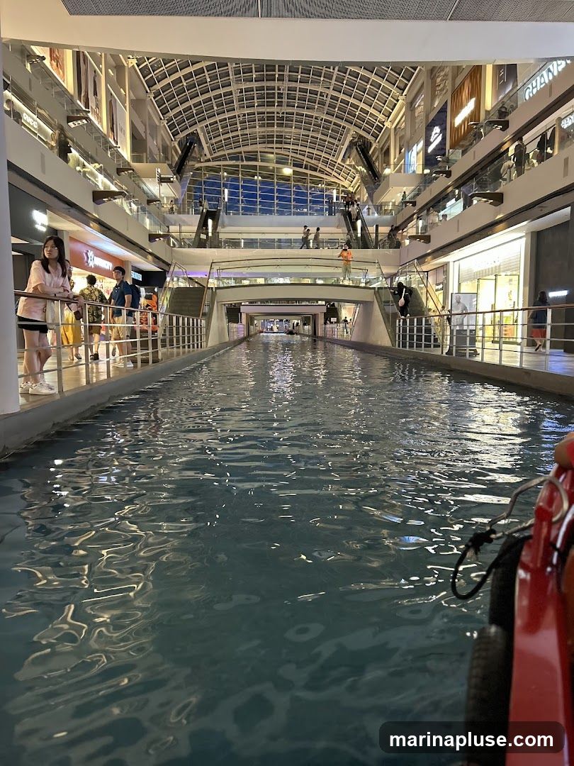 A view of an indoor canal running through a multi-level shopping mall. People are seen walking on elevated walkways and escalators. The canal reflects the lights and architecture of the mall. A red boat is partially visible in the foreground.