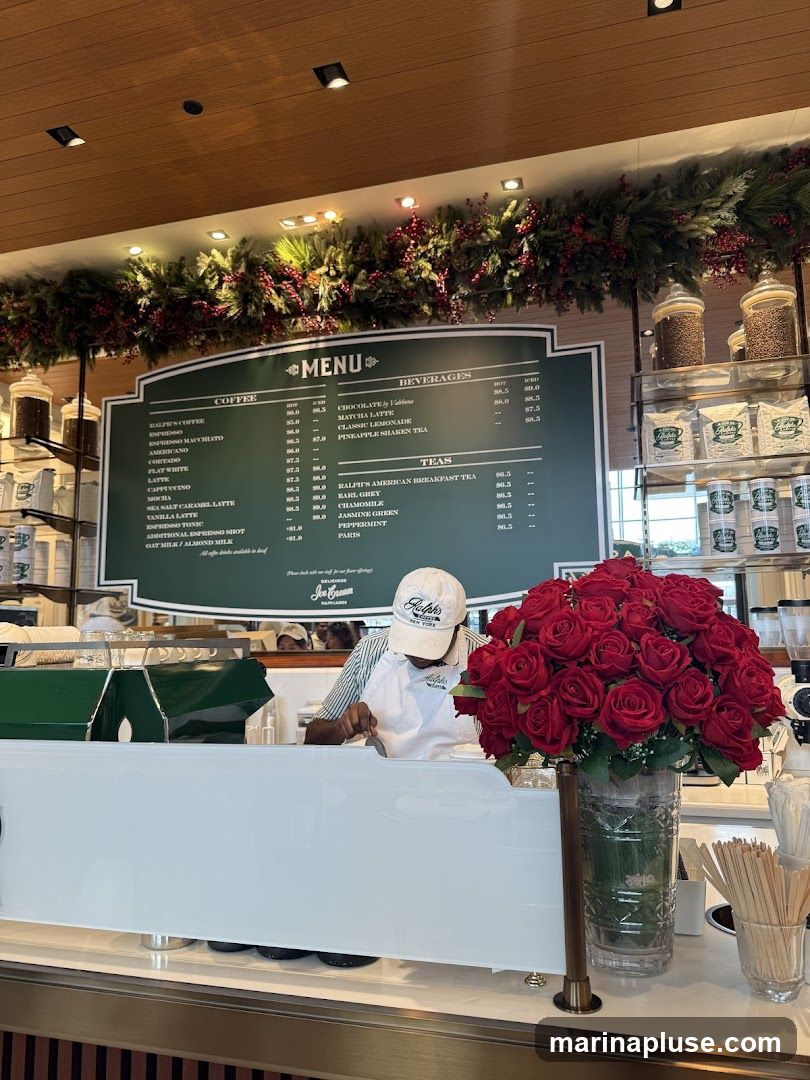 A menu board displays coffee, beverages, and teas. A barista is visible behind the counter, and a vase of red roses is in the foreground.