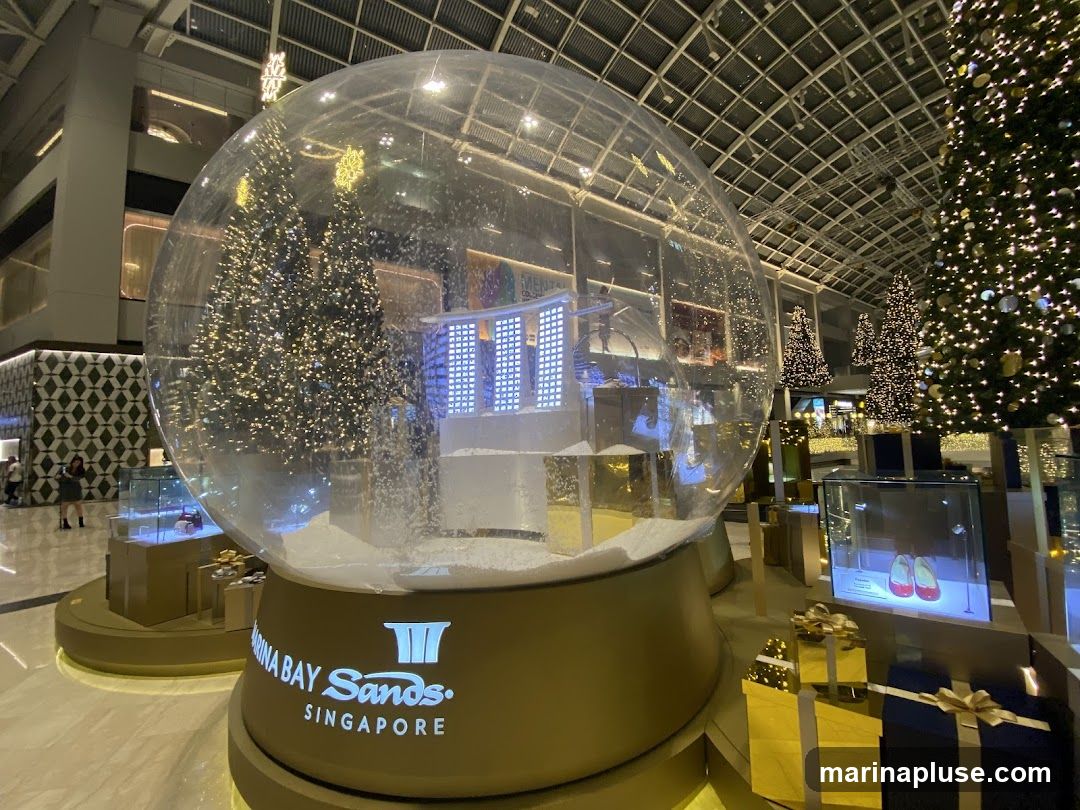 A large snow globe with a miniature cityscape and Christmas trees inside, sits on a golden base with the "Marina Bay Sands Singapore" logo. The globe is surrounded by decorated Christmas trees, gift boxes, and display cases. The scene is set indoors, likely in a shopping mall, with a high, glass-paneled ceiling.