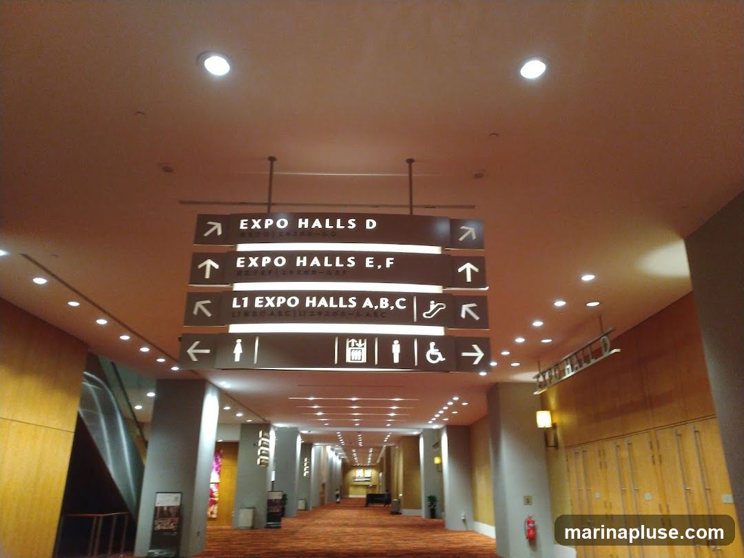 A long hallway with directional signs hanging from the ceiling. The signs indicate directions to various expo halls and include icons for restrooms and accessibility. The hallway has carpeted floors, wooden paneling on the walls, and recessed lighting.