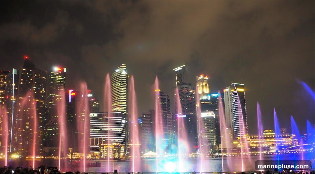 A nighttime view of a fountain show with colorful water jets shooting up against a backdrop of illuminated skyscrapers. The water is lit with pink, blue, and green lights, creating a vibrant spectacle. Silhouettes of people can be seen in the foreground watching the show. The city skyline is visible behind the fountains, with many buildings lit up.
