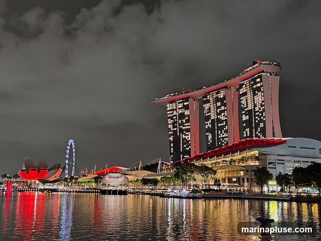 A nighttime view of the Marina Bay Sands complex in Singapore, with its iconic three towers and sky park illuminated. The ArtScience Museum, Singapore Flyer, and other waterfront structures are also visible. The scene is reflected in the calm waters of the bay.