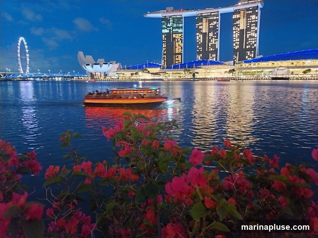 A scenic view of the Singapore skyline at dusk, featuring the Marina Bay Sands hotel, the ArtScience Museum, and the Singapore Flyer. A tour boat cruises across the water, illuminated by its lights and reflecting on the rippling surface. Pink bougainvillea flowers in the foreground add a touch of color to the twilight scene.