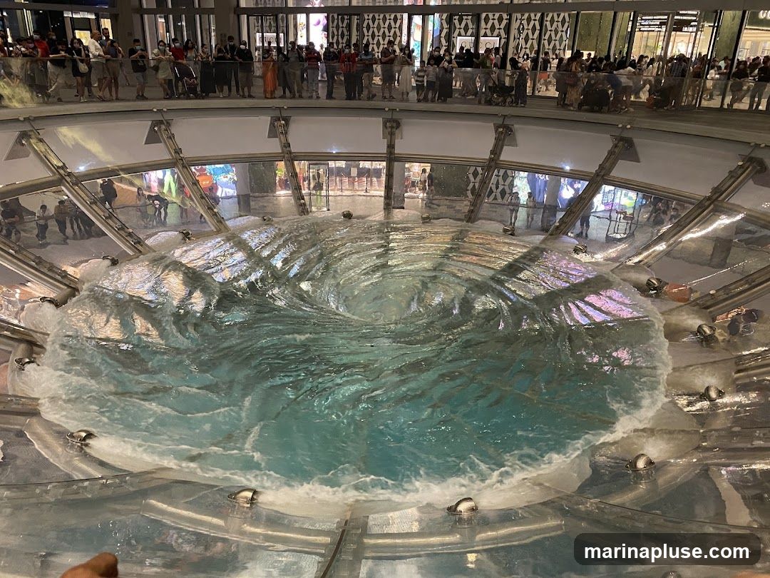 A large, circular indoor fountain creates a vortex effect with swirling turquoise water. People are gathered on an upper level, observing the spectacle. The surrounding architecture suggests a modern shopping mall or public space with reflective surfaces and bright lighting.
