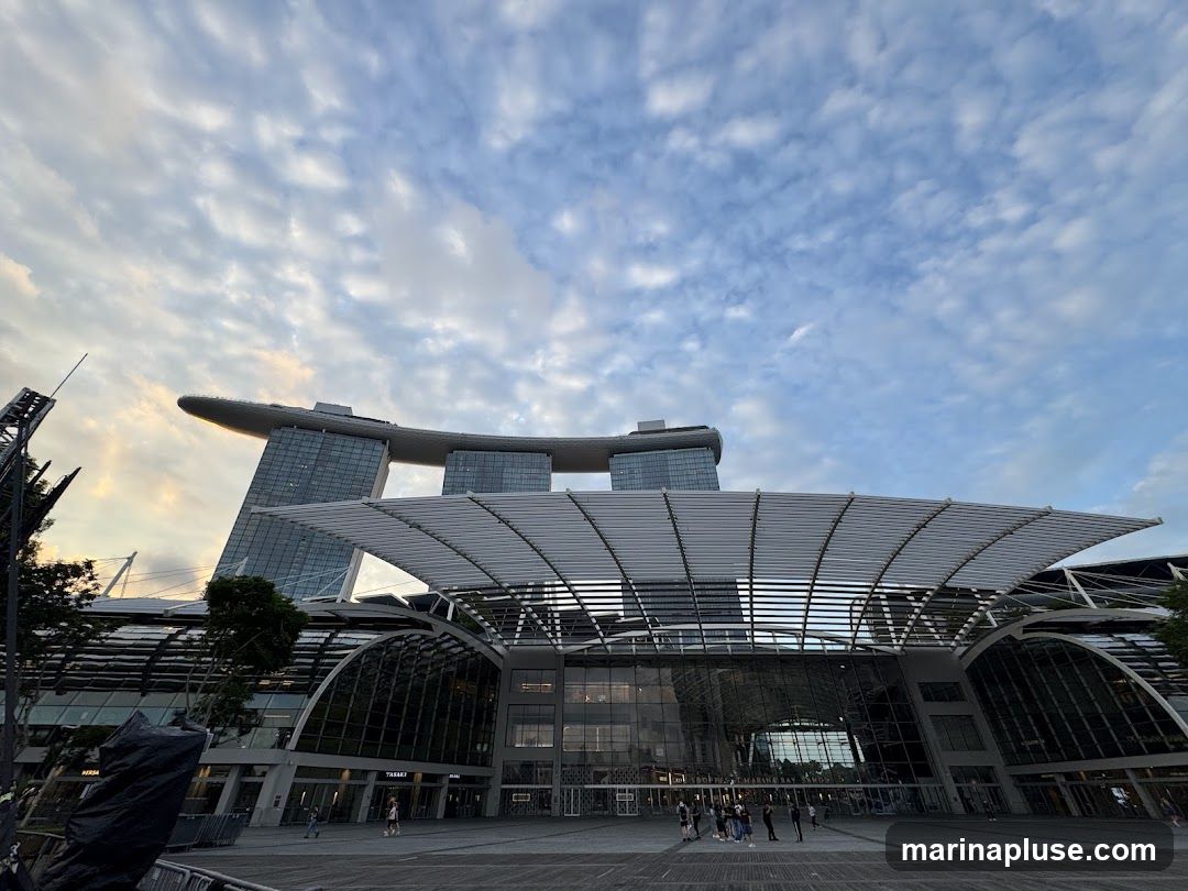 A wide-angle shot of the Marina Bay Sands complex in Singapore, featuring the iconic three towers topped with a ship-like structure. In the foreground, the expansive roof of the convention center is visible, with its distinctive ribbed design. The sky above is filled with scattered clouds, suggesting either early morning or late afternoon light. Several people are seen walking in the plaza area in front of the building.
