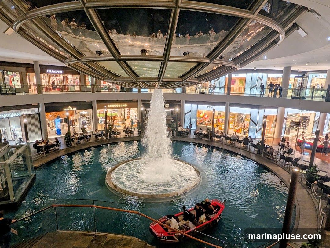 An indoor shopping mall features a large water feature with a fountain in the center. A small boat with people is navigating the water. Surrounding the water are multiple levels of shops, including visible storefronts for Armani, Stone Island, Longchamp, and Fendi. People are seen dining at tables near the water and browsing in the shops. The ceiling has a reflective surface, showing more people and the architecture above.