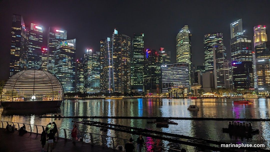 A panoramic view of the Singapore skyline at night, featuring numerous illuminated skyscrapers and a distinctive domed building on the waterfront. The reflections of the city lights shimmer on the water. Several boats are visible on the bay, and people are seen walking along the promenade in the foreground.