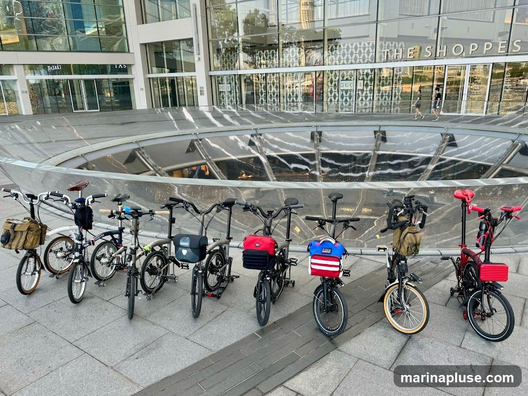 A row of seven bicycles, some folded and some unfolded, are parked on a paved walkway in front of a modern building with large glass windows. The building has signage that reads "THE SHOPPES" and "TASAKI". Some of the bicycles have bags or baskets attached.