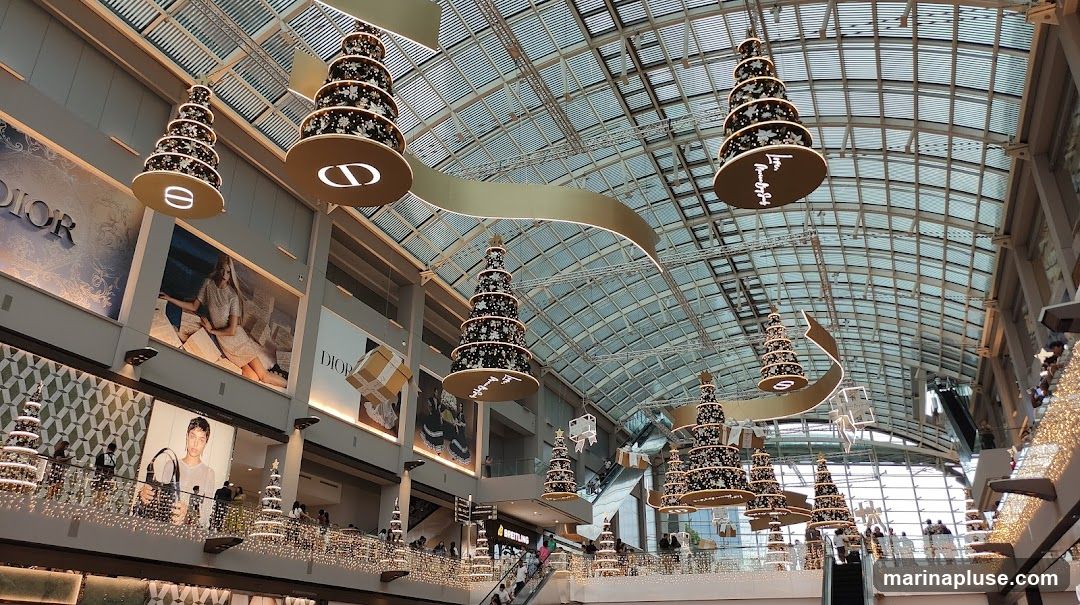 A wide shot of a mall interior decorated for Christmas. Large, tiered Christmas tree-shaped ornaments hang from the ceiling, adorned with lights and snowflakes. Banners with the Dior logo and fashion imagery are visible on the walls. Escalators and shoppers can be seen on multiple levels, with festive string lights lining the railings. The ceiling is a glass and metal structure, allowing natural light to filter in.