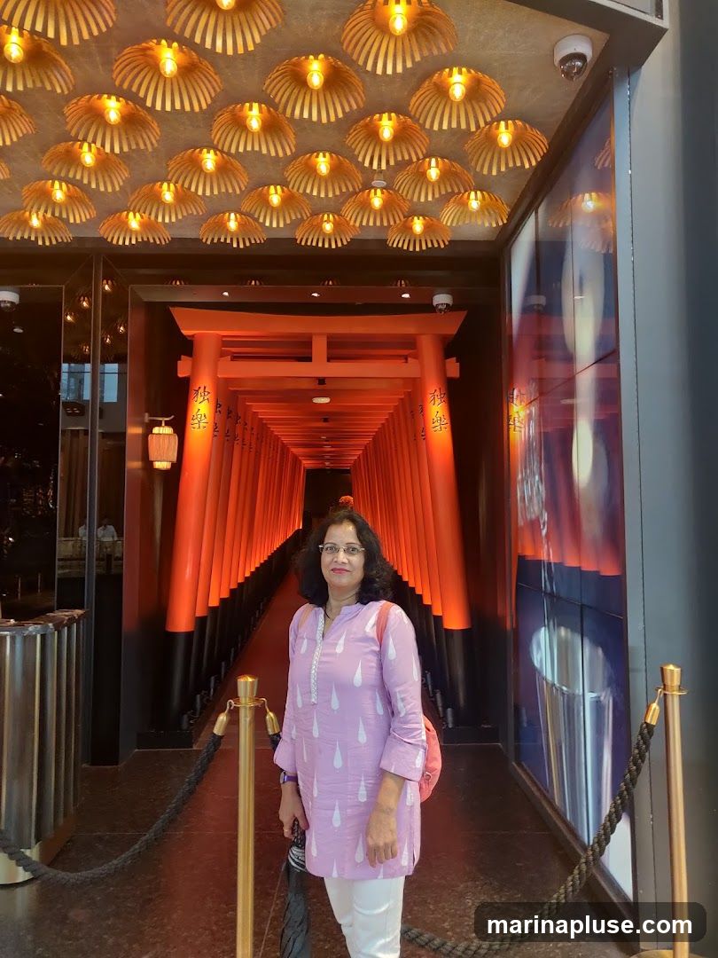 A woman stands in a hallway decorated with traditional Japanese torii gates and overhead bamboo-style lamps. The torii gates are painted a vibrant orange and have Japanese characters on them. The lamps have a unique, fan-like design. The woman is wearing a light purple tunic with white patterns and white pants. She is holding an umbrella and has a pink backpack. The corridor appears to be an entrance or walkway within a building, possibly a restaurant or entertainment venue.