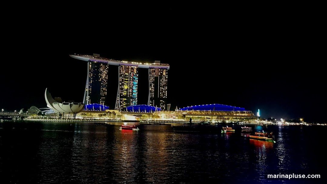 A nighttime view of the Marina Bay Sands complex in Singapore, illuminated against a dark sky. The iconic three towers are visible, with lights twinkling from many windows. In front of the towers, a building with a blue roof is lit up, and a distinctive lotus-shaped structure is also illuminated. Several boats are on the water in the foreground, reflecting the lights of the city.