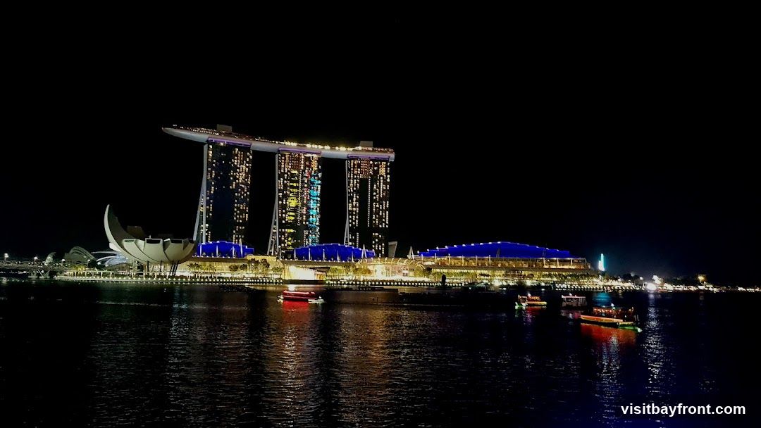 A nighttime view of the Marina Bay Sands complex in Singapore, illuminated against a dark sky. The iconic three towers are visible, with lights twinkling from many windows. In front of the towers, a building with a blue roof is lit up, and a distinctive lotus-shaped structure is also illuminated. Several boats are on the water in the foreground, reflecting the lights of the city.