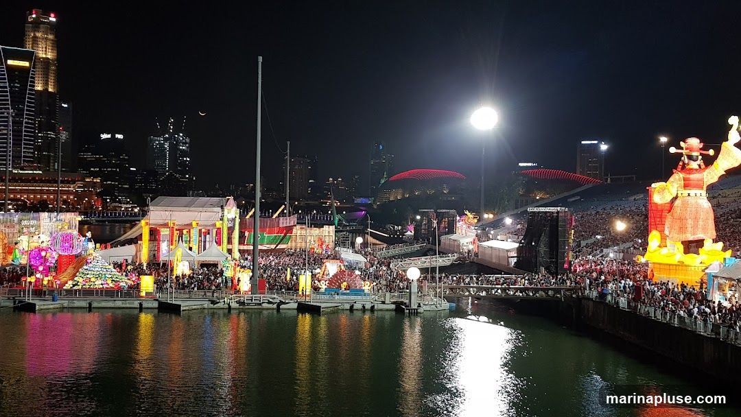 A vibrant nighttime scene of a festival, likely celebrating Chinese New Year, with numerous illuminated lanterns and large decorative figures. The festival is set along a waterfront, with a cityscape visible in the background, including tall buildings and a distinctive domed structure. A large crowd of people is gathered, enjoying the festivities. The water in the foreground reflects the colorful lights of the event.