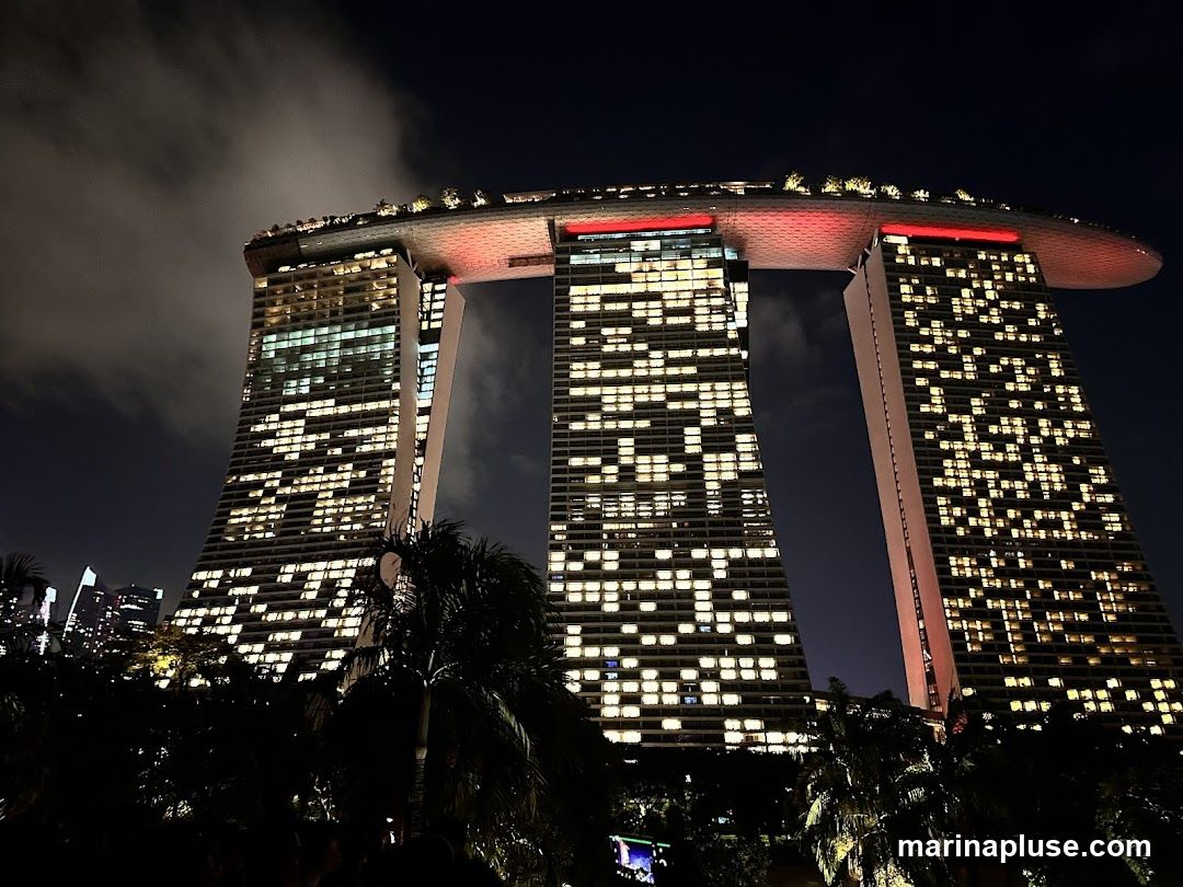 A nighttime view of the Marina Bay Sands hotel in Singapore, with its iconic three towers connected by a sky park. The building is illuminated, with many windows lit up, creating a grid pattern against the dark sky. Palm trees and other foliage are visible in the foreground, silhouetted against the bright lights of the hotel.