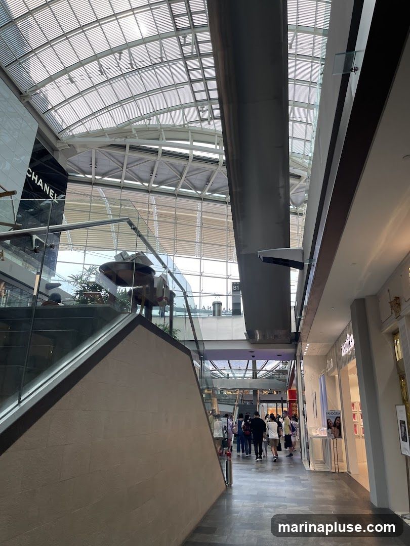 An interior view of a modern shopping mall with a glass ceiling. A staircase with glass railings is on the left, leading up to a CHANEL store. A walkway with shops on either side is visible in the distance, with several people walking. The Pandora store is visible on the right.