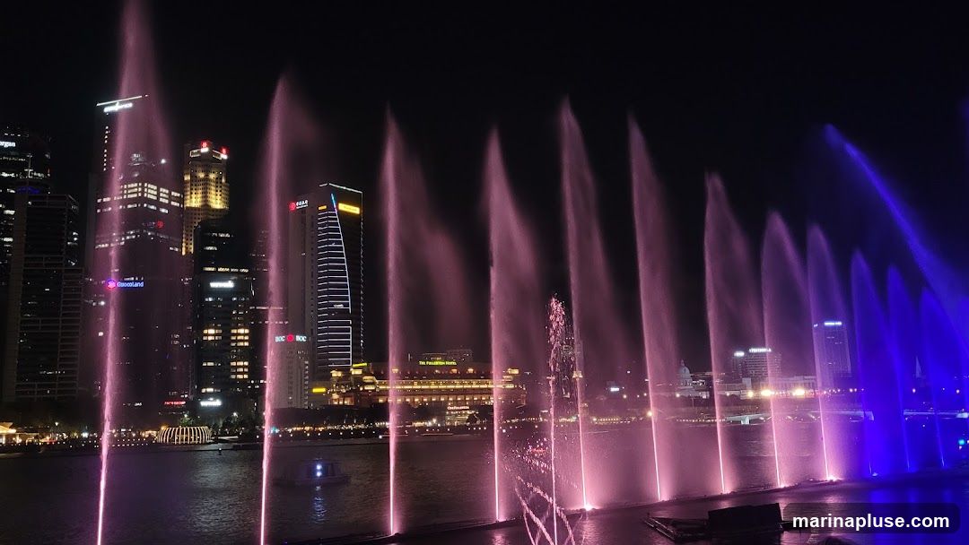 A nighttime view of a water fountain show with colorful lights, set against a backdrop of a modern city skyline. The fountains spray water high into the air, illuminated by pink and blue lights. Several tall buildings with illuminated windows are visible in the background, reflecting the city lights. A body of water is in the foreground, with a few boats.