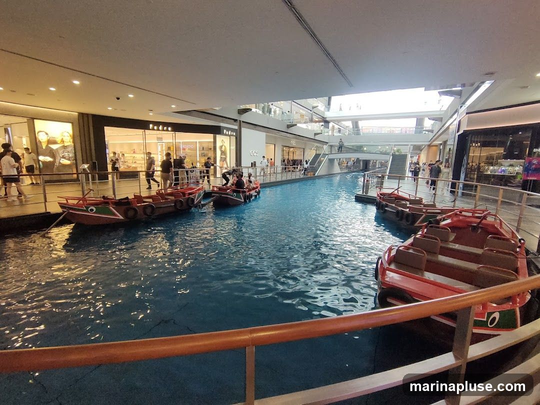An indoor canal with several small boats docked along the sides. People are visible walking along the walkways next to the canal, and shops are visible in the background. The canal appears to be part of a shopping mall or entertainment complex.