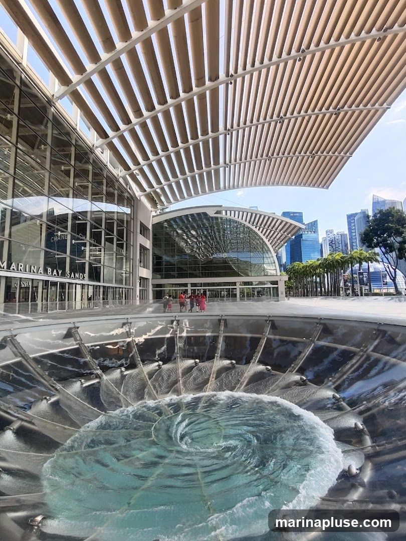 A wide shot of the exterior of Marina Bay Sands in Singapore, featuring the building's modern architecture and a large, swirling water feature in the foreground. The sky is clear blue, and the city skyline is visible in the background.