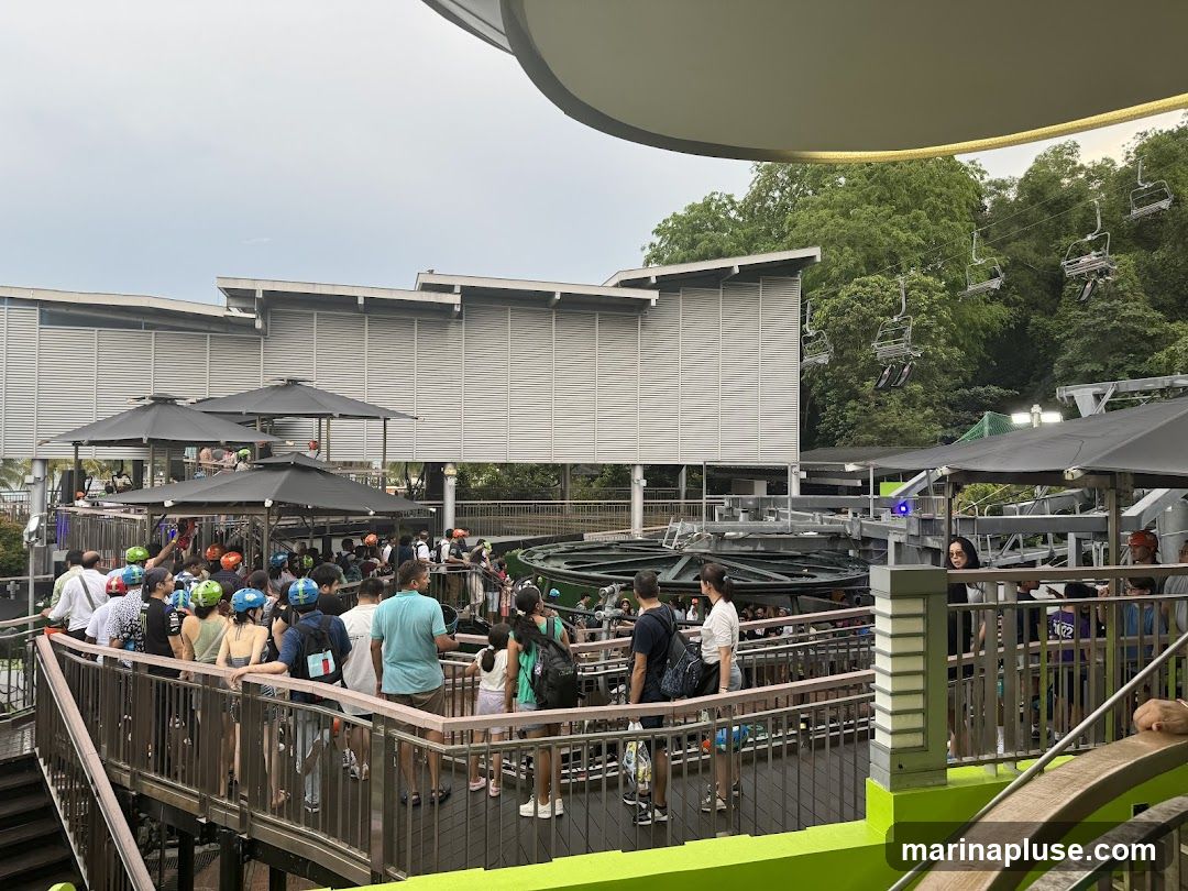 A crowd of people are waiting in line on a raised walkway, many wearing helmets, for a cable car ride. The cable cars are visible in the background, suspended from wires above a lush green treeline. The walkway has metal railings and is bordered by green structures. A modern building with horizontal louvers is visible behind the waiting area.