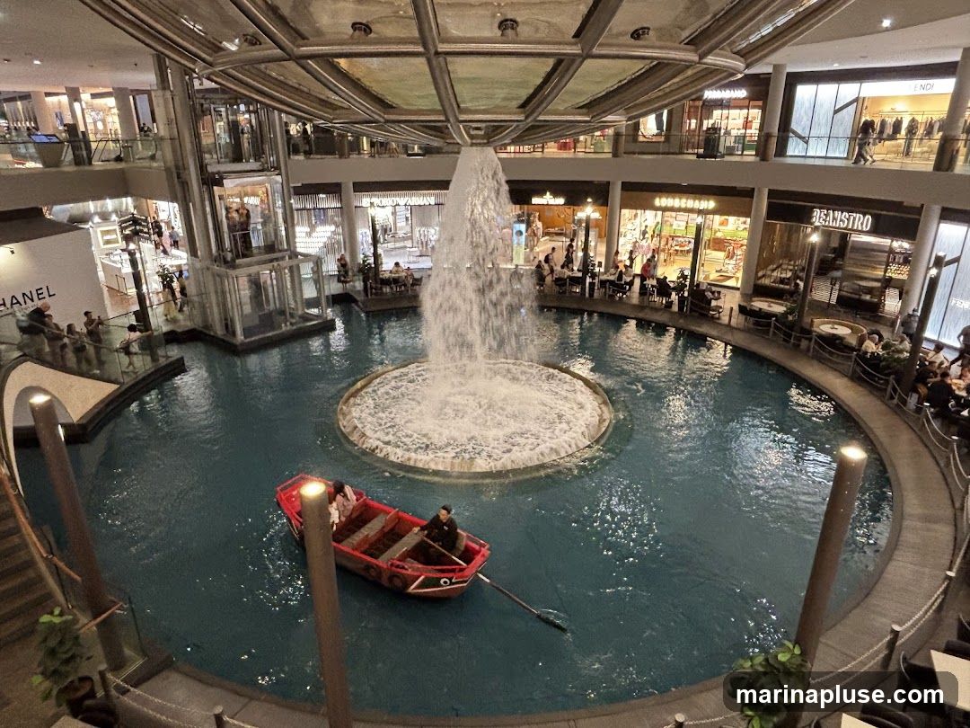 An indoor shopping mall features a circular canal with a large fountain in the center. A small red boat with people is being rowed through the water. Surrounding the canal are multiple levels of shops and restaurants, with visible signage for brands like CHANEL, LONGCHAMP, and FENDI. People are seen walking, dining, and observing the fountain.