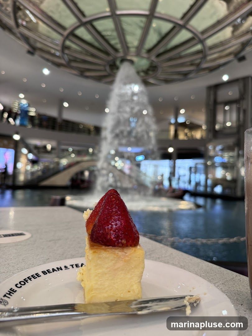 A slice of cheesecake topped with strawberries sits on a plate with a fork. In the background, a large indoor fountain cascades water under a domed ceiling. The setting appears to be a shopping mall or a similar public space with multiple levels visible. The plate has the text "THE COFFEE BEAN & TEA" printed on it.