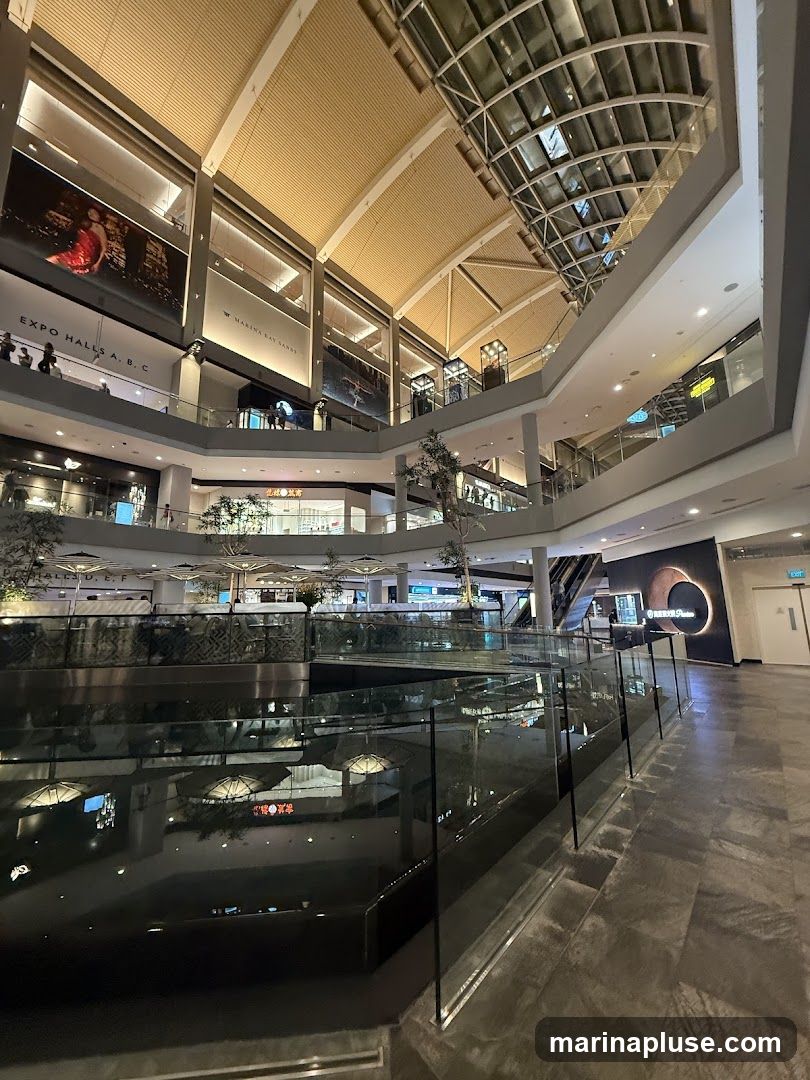The image shows the interior of a multi-level shopping mall. A large, dark water feature is in the foreground, reflecting the lights and architecture of the building. The upper levels of the mall are visible, with shops, escalators, and large display screens. The ceiling is high and features a complex glass and metal structure. Signage for "MARINA BAY SANDS" is visible on one of the upper levels.