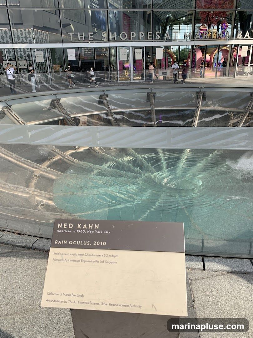 A view of the exterior of The Shoppes at Marina Bay Sands, a large shopping mall. In the foreground is a plaque describing the "Rain Oculus, 2010" sculpture by Ned Kahn. The sculpture itself, made of stainless steel, acrylic, and water, is visible in a circular pool. People are seen walking in front of the mall entrance in the background.