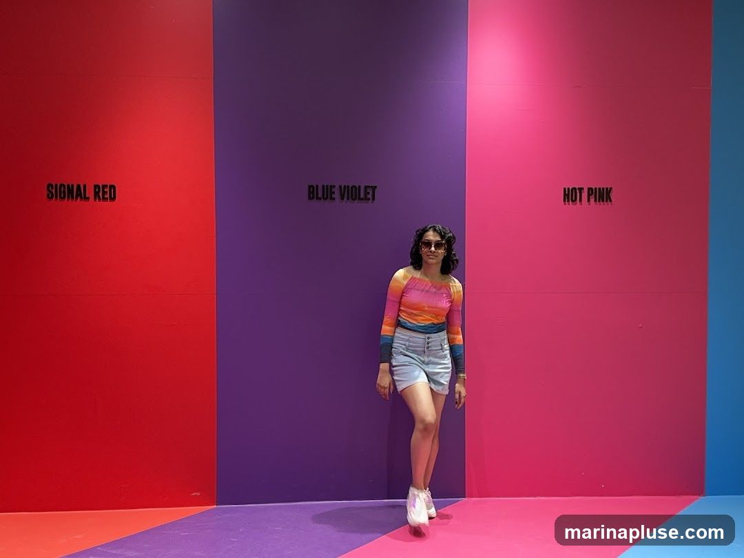 A woman stands in front of a wall divided into sections of bright colors: red, purple, and pink. Each color section has a label above it: "SIGNAL RED", "BLUE VIOLET", and "HOT PINK". The floor also features a colorful geometric pattern. The woman is wearing a striped off-the-shoulder top, denim shorts, sunglasses, and white sneakers.
