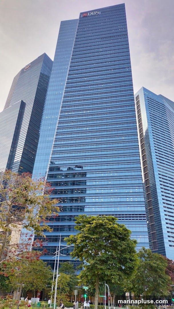 A low-angle shot of a modern, glass-facade skyscraper with the DBS logo visible at the top. The building is surrounded by other tall buildings and lush green trees in the foreground. The sky is overcast.
