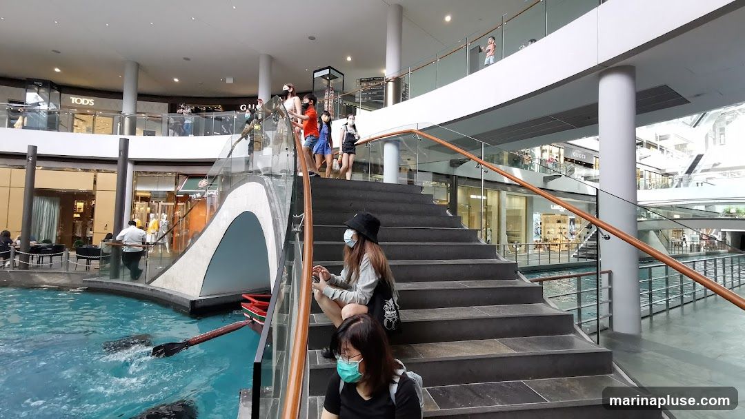 A group of people are walking up and sitting on a wide staircase in a modern shopping mall. Below the stairs is a canal with a small boat. Shops are visible on multiple levels of the mall, including one with a "TOD'S" sign and another with a "GUCCI" sign.
